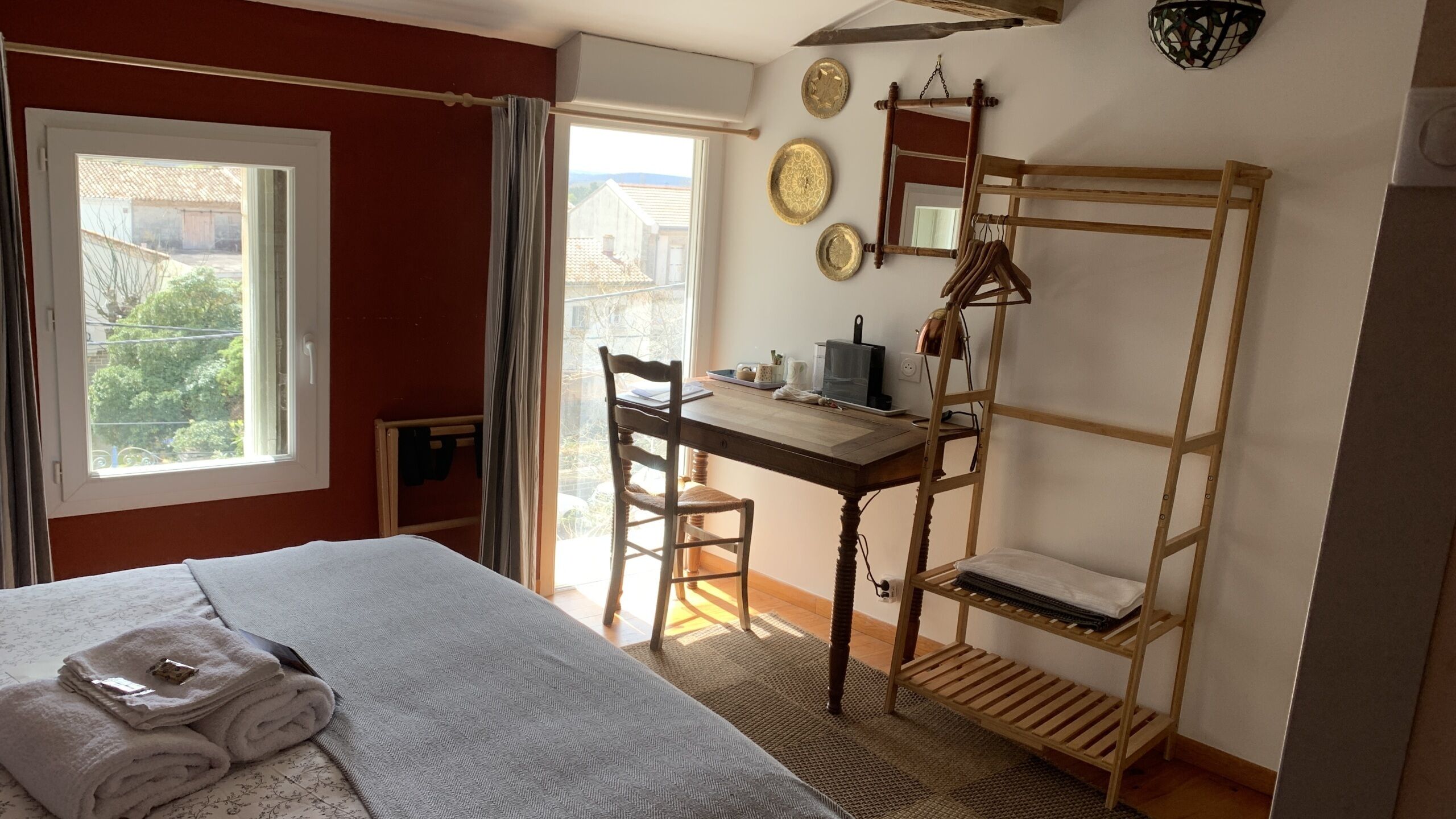 Una habitación con cama, escritorio, silla, escalera de madera y ventana con vistas a edificios y árboles.