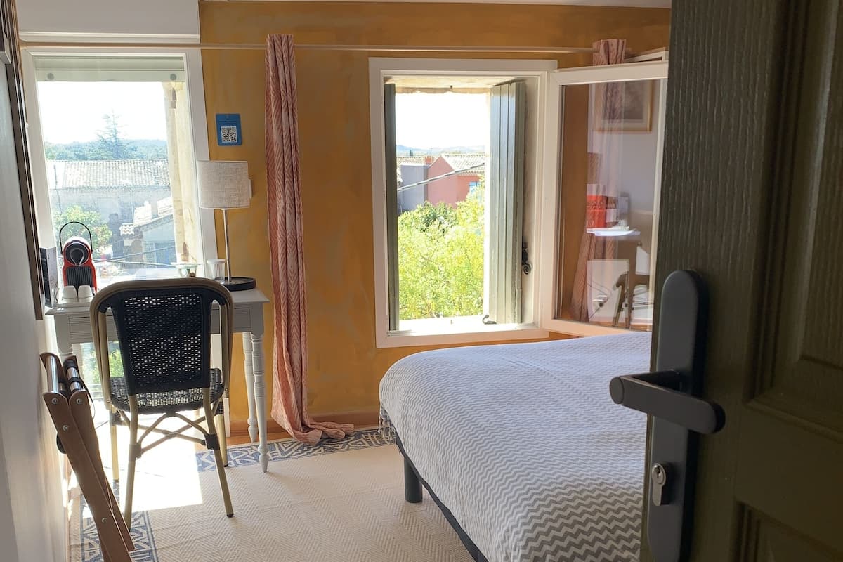 A bedroom with a bed, a desk, and a chair. There is a window with a view of greenery outside.