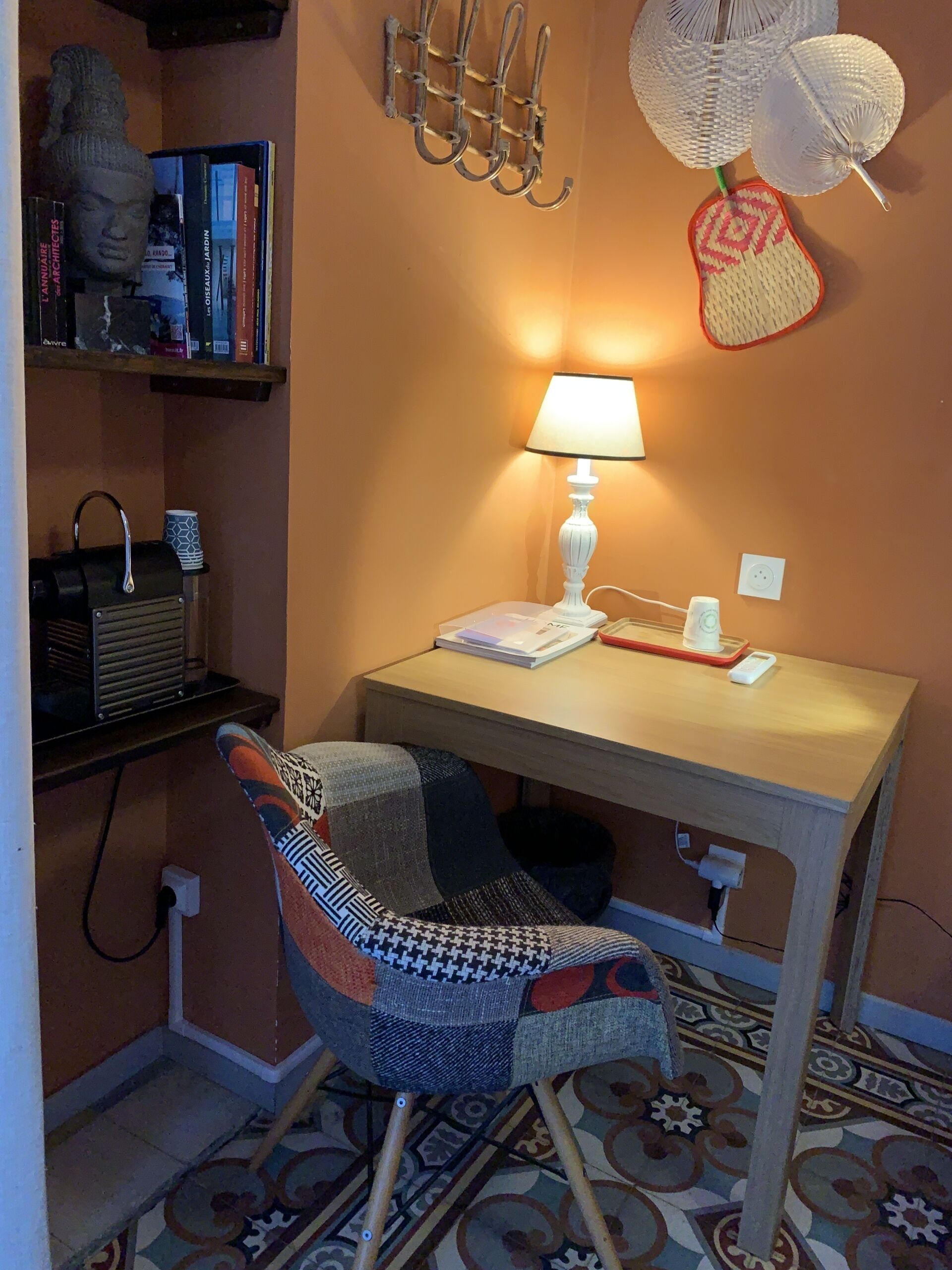 Um quarto pequeno e acolhedor com uma mesa de madeira, uma poltrona estampada, uma luminária, uma estante e um objeto decorativo na parede.