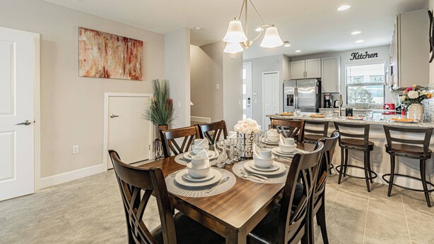 A dining area with a wooden table set for six, a kitchen in the background, and a painting on the wall.