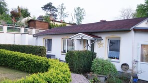 Ein eingeschossiges Haus mit dunklem Dach, weißen Außenwänden und einem kleinen Vorgarten mit ordentlich geschnittenen Hecken und einem Blumentopf.