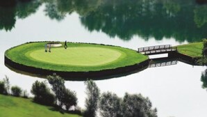 Un campo de golf con un putting green circular, un puente y un cuerpo de agua que refleja el entorno.