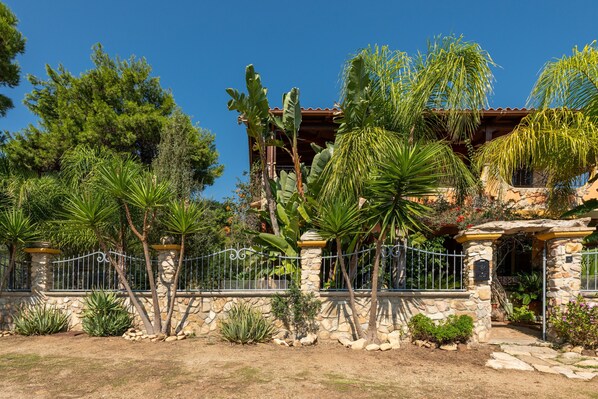 A stone-walled property with a metal gate, tropical plants, and a building with a tiled roof.