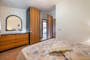 Une chambre à coucher comprenant un lit, une commode, un miroir, une armoire et donnant sur une autre pièce.