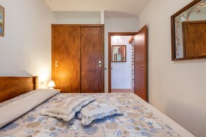 A bedroom with a bed, a wooden wardrobe, a mirror, and a door leading to another room.