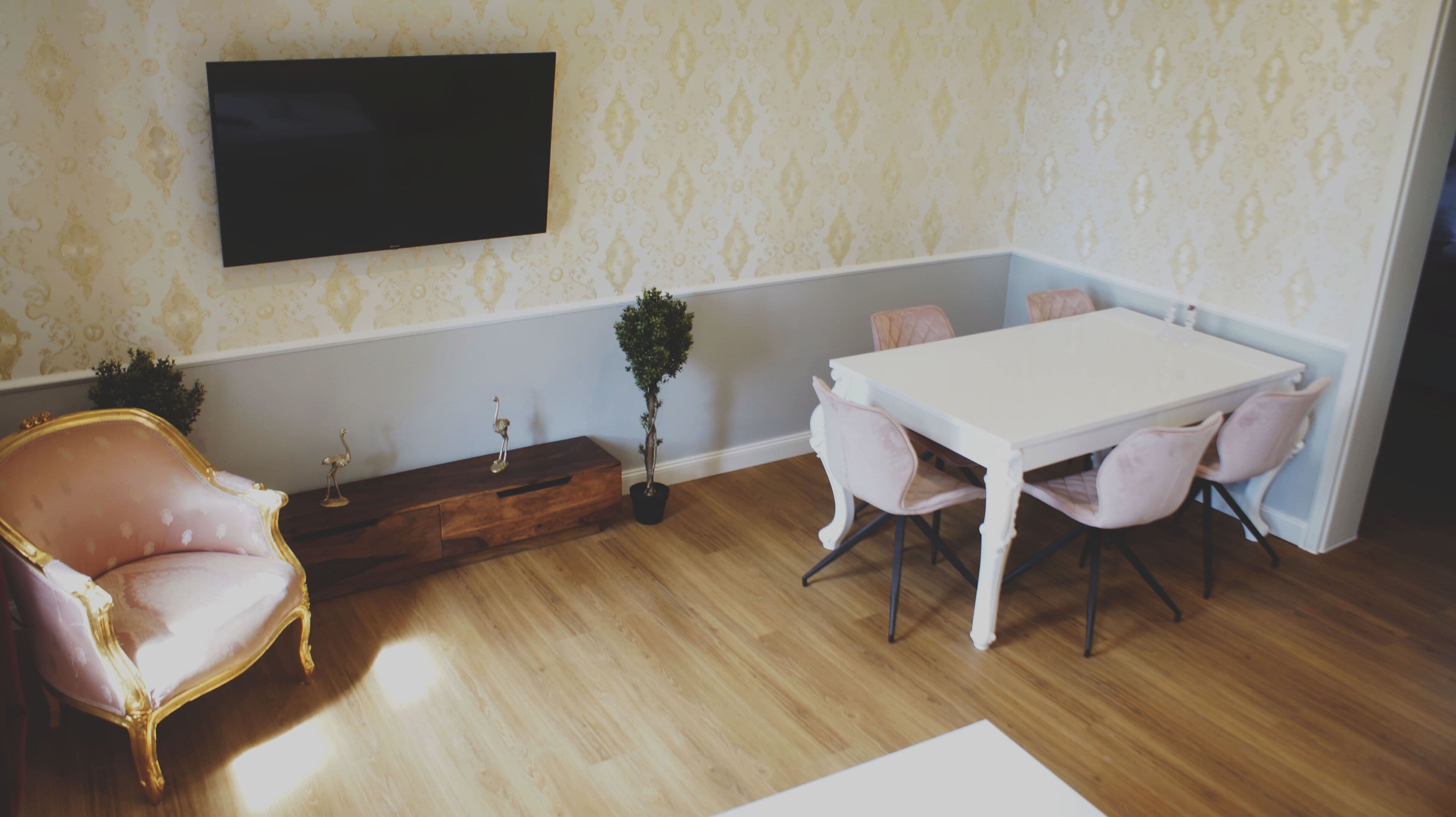 En matplats med ett vitt bord och rosa stolar, en fåtölj med guldfärgad ram, en krukväxt och en platt-TV som är monterad på väggen.