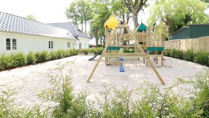 Un parque infantil con tobogán y estructuras para escalar, rodeado de arena y vegetación, con una casa al fondo.