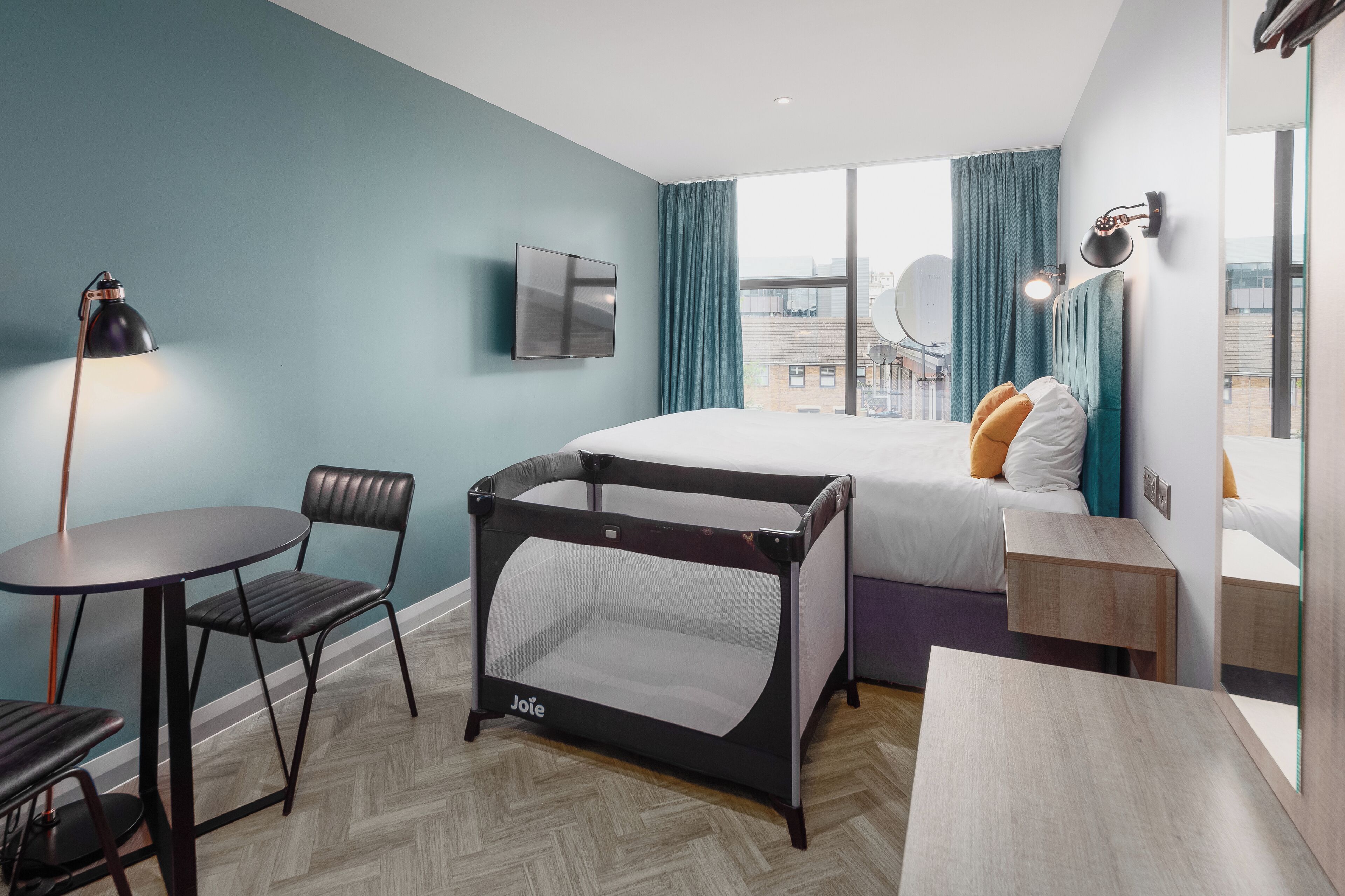 A modern hotel room with a bed, a desk, a chair, a table, a TV, and a window with curtains.