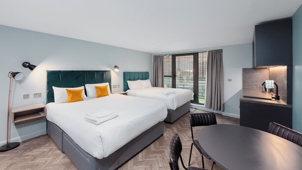 A modern hotel room with two beds, a dining table, a kitchenette, and a large window with curtains. - Aptel East (London)