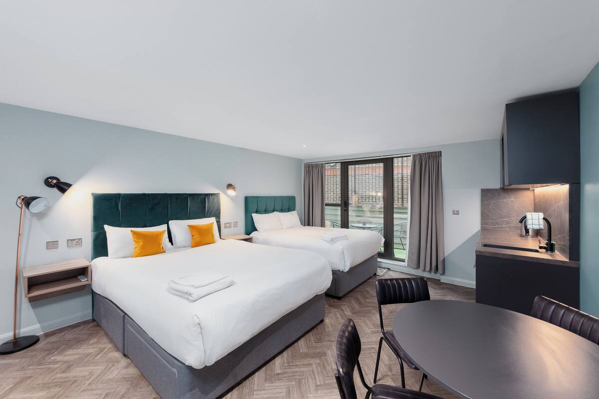 A modern hotel room with two beds, a dining table, and a kitchenette.
