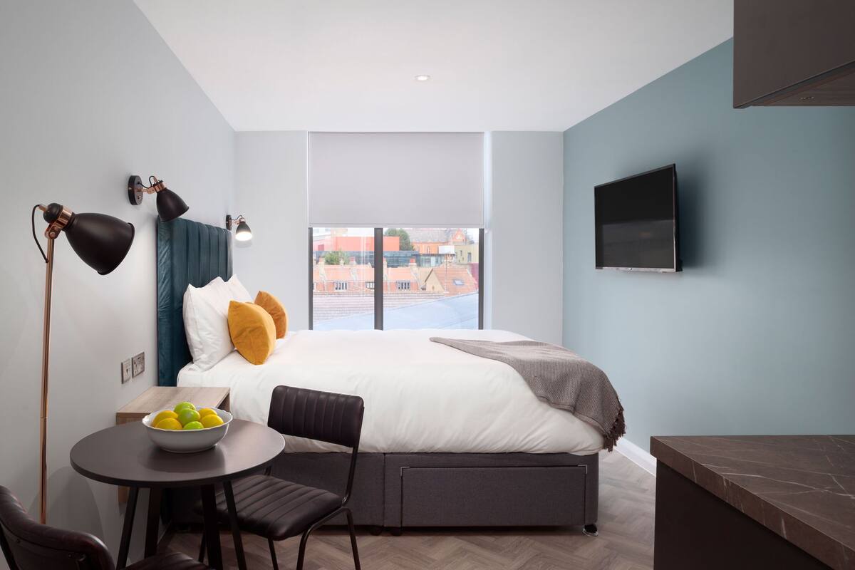 A modern hotel room with a large bed, a flat-screen TV, a dining table with a bowl of fruit, and a view of the cityscape through the window.