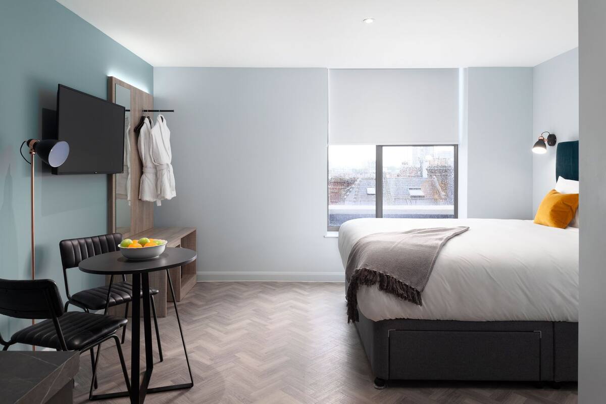 A modern hotel room with a bed, a desk with a chair, a television, and a large window.