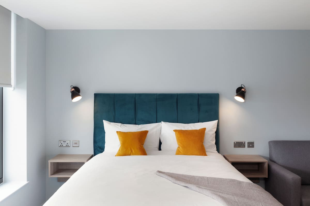 A modern hotel room with a teal headboard, white bedding, and two bedside tables with lamps.