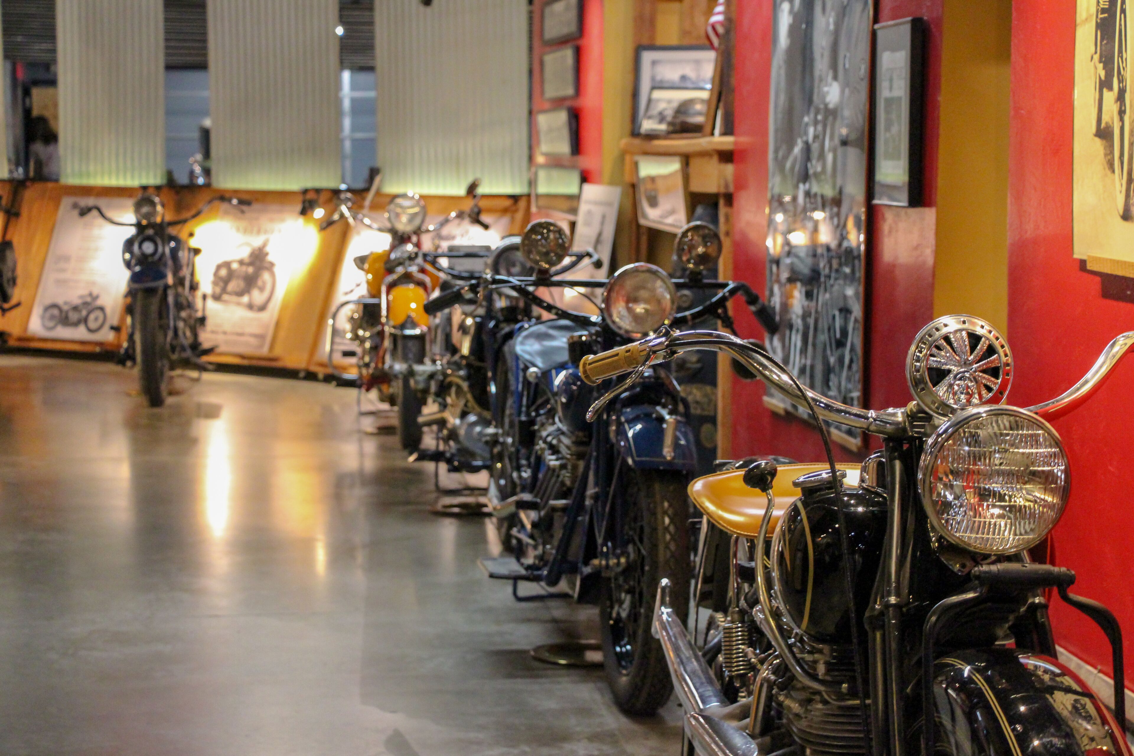 Une rangée de motos anciennes exposées dans un musée ou une salle d’exposition, avec des murs rouges et des tableaux encadrés accrochés aux murs.