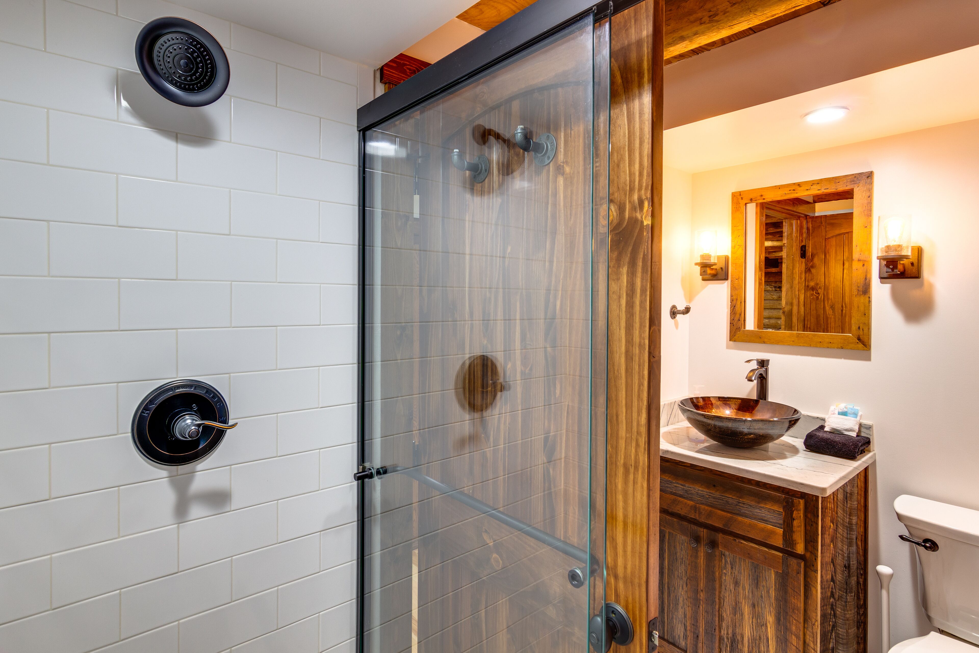 A modern bathroom with a glass shower enclosure, a wooden vanity, and a white tiled wall.