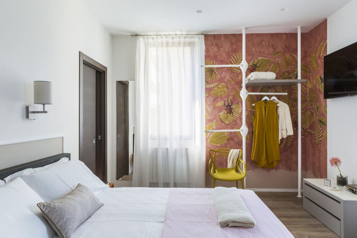 Une chambre d’hôtel moderne avec un lit bien fait, une armoire où sont suspendus des vêtements, un téléviseur fixé au mur et une fenêtre avec des rideaux légers.