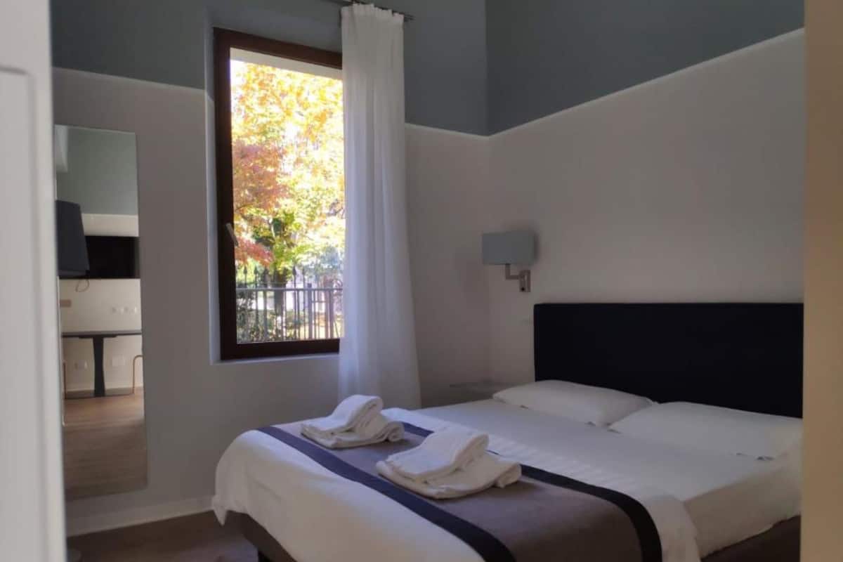 Une chambre d’hôtel avec un lit, une fenêtre avec des rideaux, une table de chevet et une lampe fixée au mur.