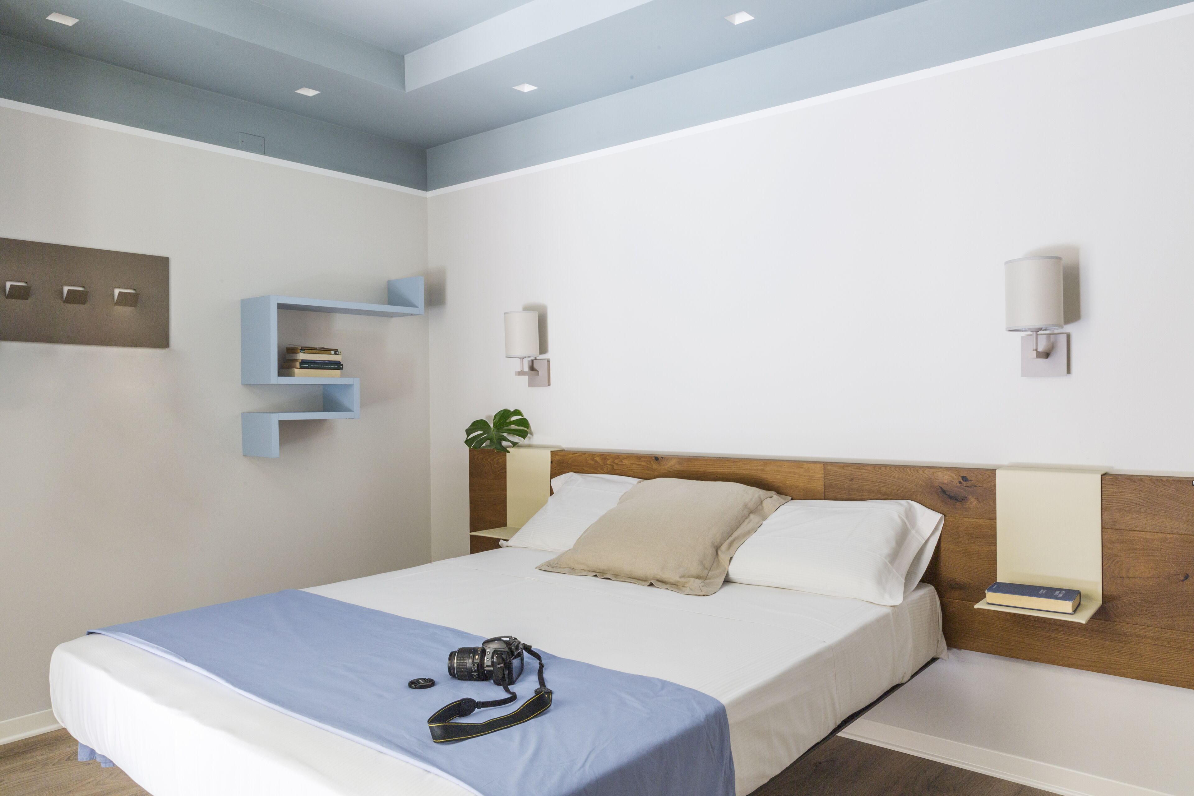 Una cama bien hecha con colcha azul, una cámara sobre la cama y un cabecero de madera. Hay dos lámparas de pared y un estante pequeño con libros.