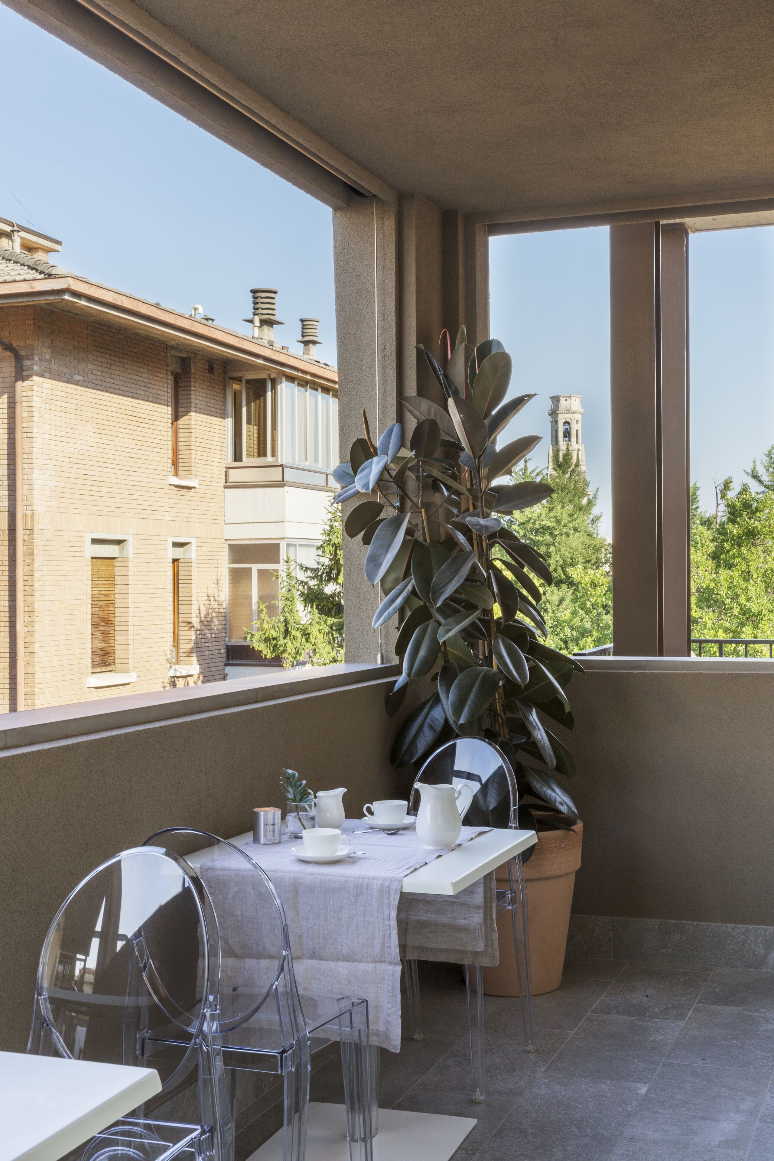 Ein Balkon mit einem für zwei Personen gedeckten Tisch, transparenten Stühlen, einem Topf mit einer Pflanze und Blick auf Gebäude und Bäume.
