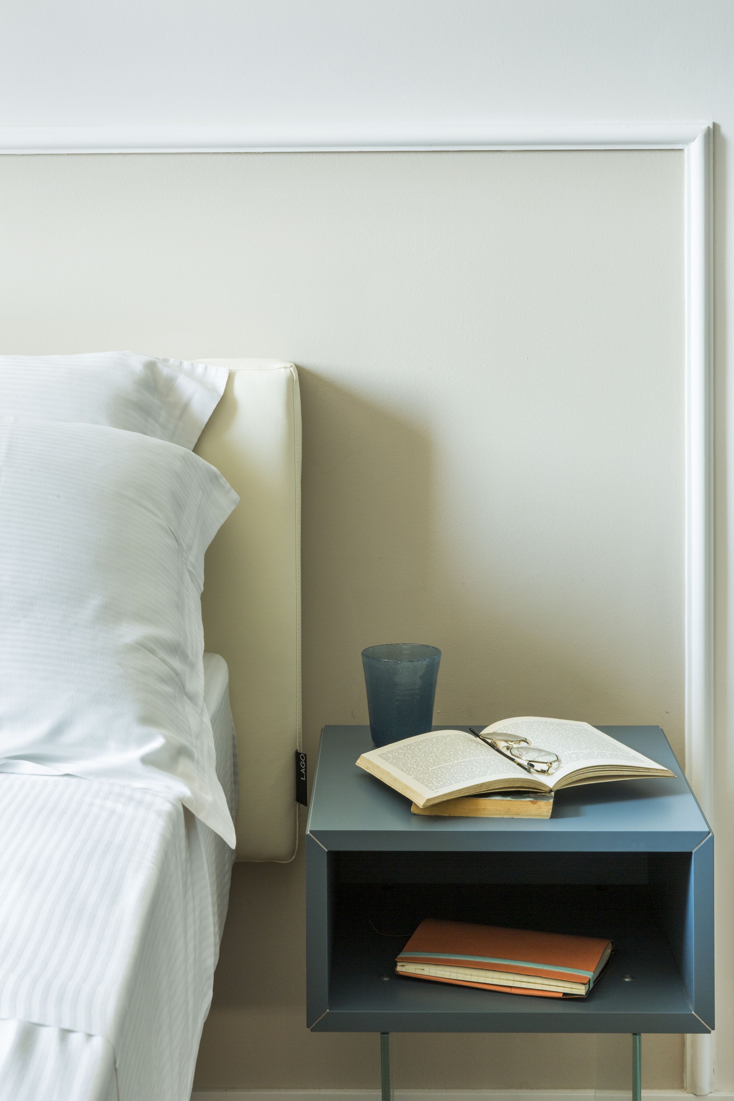 Una cama bien tendida con sábanas blancas, una mesita de noche con un libro y gafas, y una lámpara.