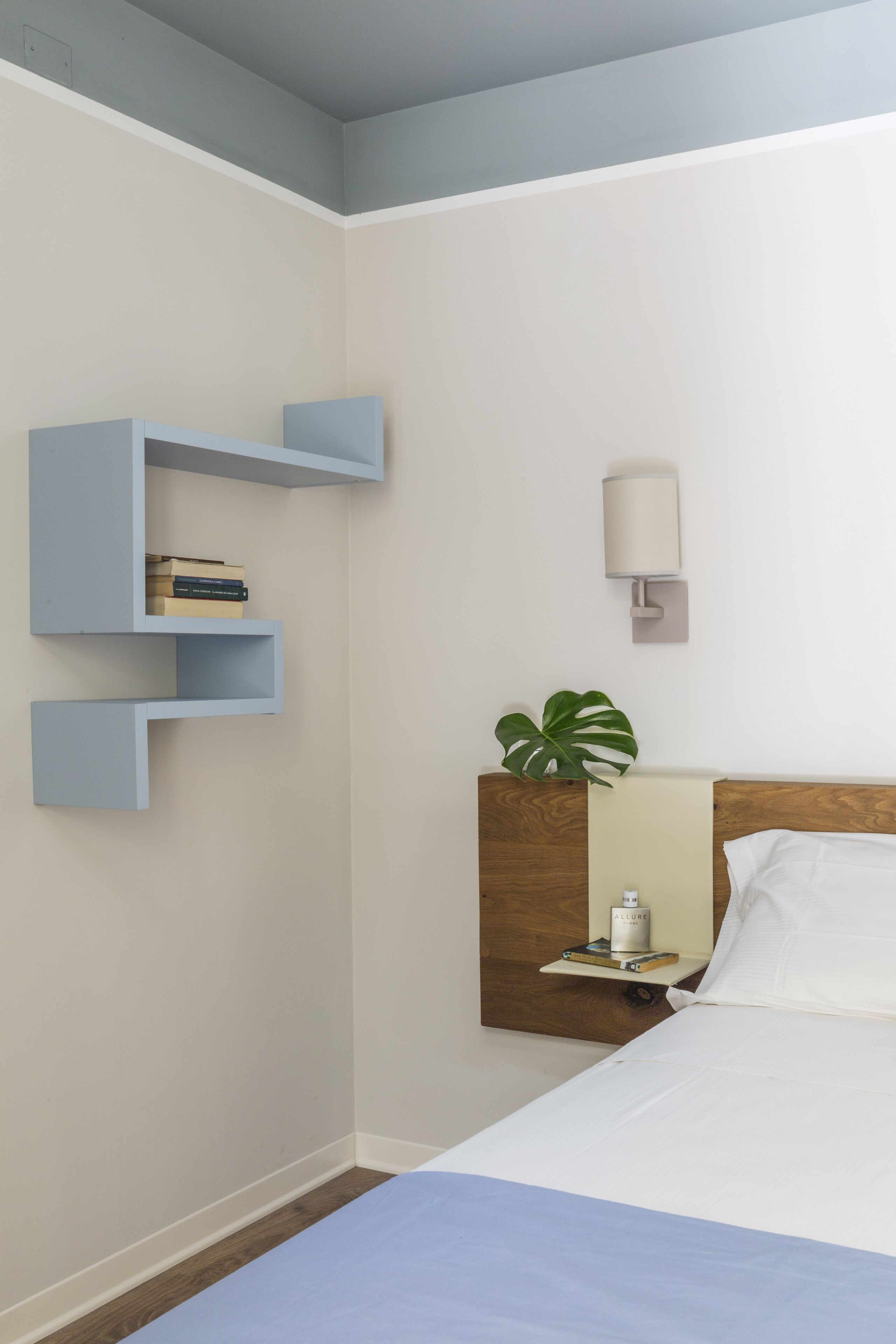 A hotel room with a bed, a nightstand, a wall-mounted shelf with books, a wall-mounted lamp, and a potted plant.
