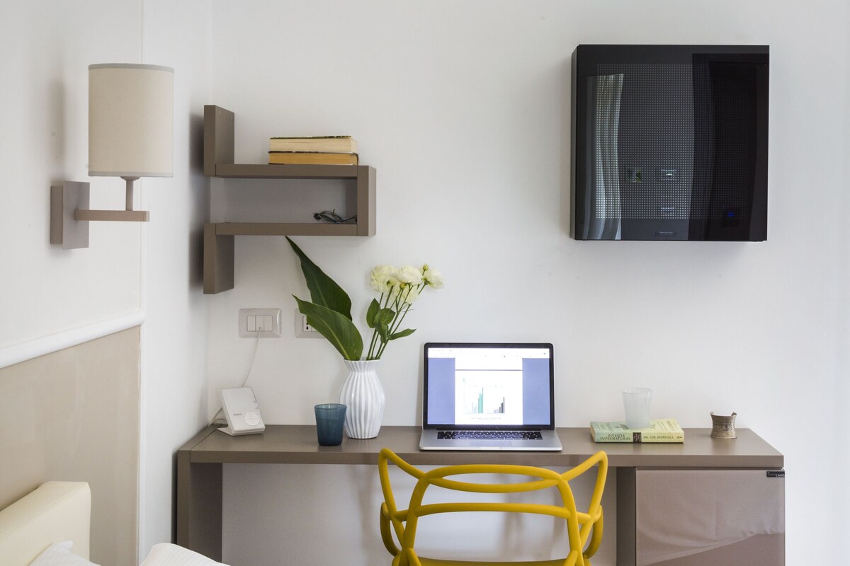 Habitación de hotel con cama, escritorio con computadora portátil, silla, televisor y estante en la pared con libros y un florero.