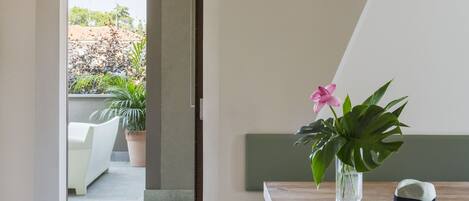 A modern interior with a wooden desk, a clear glass vase with a pink flower, and a view of an outdoor patio with plants and a white chair.