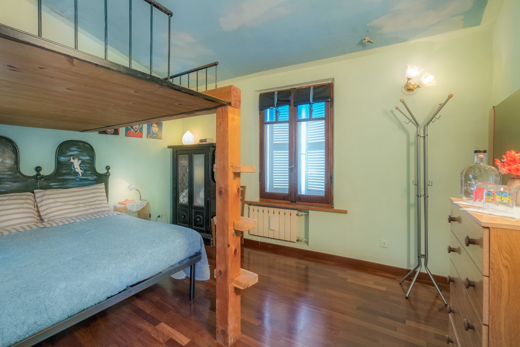 Un dormitorio con literas, una ventana con contraventanas, un radiador, una lámpara de pie y un armario con espejo.