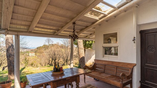 A covered patio with a wooden table and bench, a black door, and a view of trees and hills. - Entire house in the countryside with garden and sea view (Pesaro)