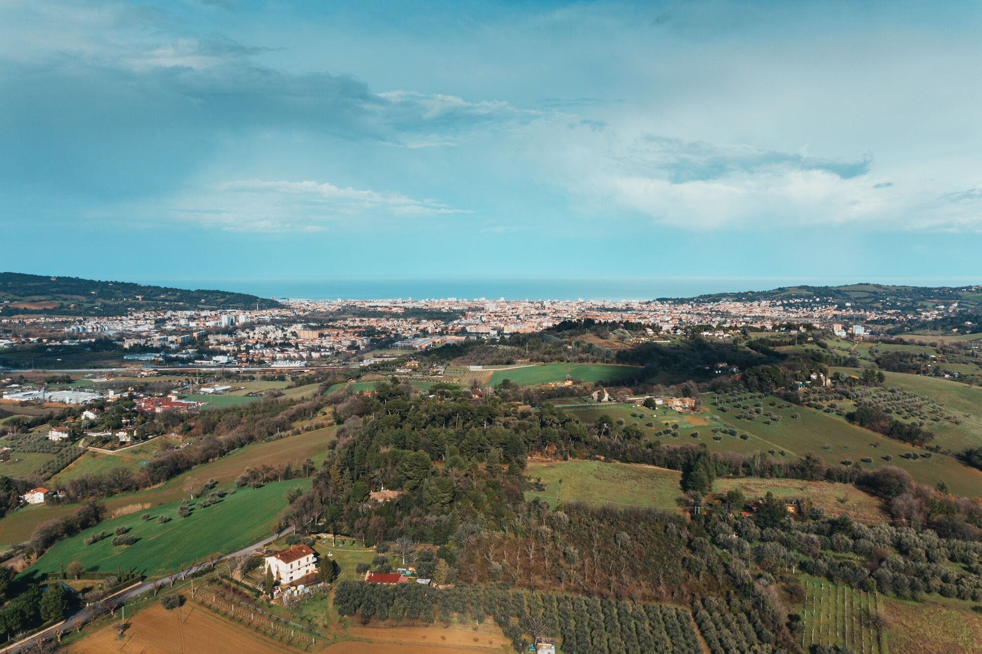 Vista aérea de uma cidade costeira, com uma mistura de paisagens urbanas e rurais, incluindo campos, árvores e uma vista distante do mar.