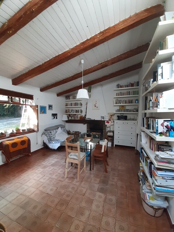 Una habitación acogedora con suelo de baldosas, vigas de madera, chimenea, una mesa de comedor con sillas y una estantería llena de libros.