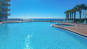 A large, clear swimming pool with lounge chairs and palm trees, surrounded by a fence and a building.