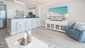 A modern kitchen with white cabinets, a central island with bar stools, a dining table with candles, and a large beach-themed painting above the sofa.