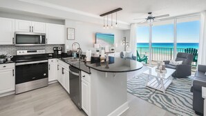 A modern kitchen with white cabinets, stainless steel appliances, and a large island. There is a dining area with a view of the ocean.