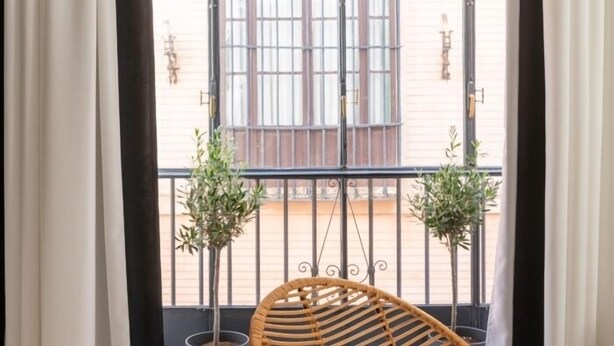 A balcony with a wicker chair, two potted plants, and a view of a building with barred windows. - Hommyhome Vera Cruz 101 (Sevilla)