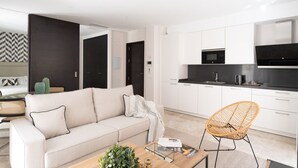 A modern living room with a sofa, a coffee table with magazines, and a wicker chair. The kitchen has white cabinets, a black countertop, and a microwave. - Hommyhome Vera Cruz 101 (Sevilla)