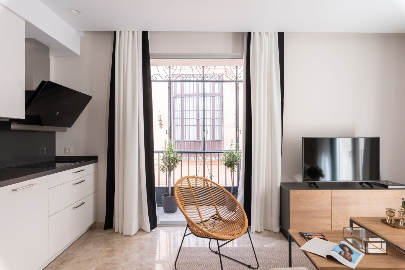 Une cuisine moderne avec un plan de travail noir, des armoires blanches et une hotte noire. Au centre de la pièce, on trouve une chaise en osier, une table à manger en bois sur laquelle sont posés un magazine et un livre, ainsi qu’un téléviseur sur un meuble en bois.