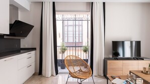 A modern kitchen with a black countertop, white cabinets, and a black range hood. There is a wicker chair in the center of the room, a wooden dining table with a magazine and a book, and a TV on a wooden stand. - Hommyhome Vera Cruz 101 (Sevilla)