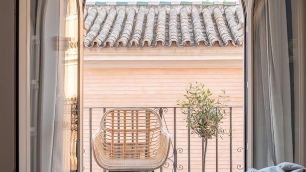 Un balcón con una silla de mimbre, una planta en maceta y vistas a un tejado tradicional con tejas.