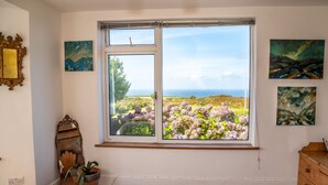 Un dormitorio con un ventanal amplio que ofrece vistas a un campo y al océano. Hay cuadros en las paredes, un aparador de madera y un espejo.