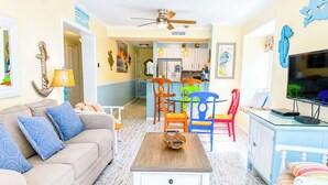 A living room with a sofa, coffee table, and chairs, a kitchen in the background, and a TV on a stand. - Experience Tilghman Beach and Racquet Club Unit 220! 3 Bedroom Ocean View Condo. Pools, Tennis, Close to Main Street! (North Myrtle Beach)