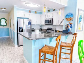 A kitchen with white cabinets, a granite countertop, a refrigerator, a microwave, and a stove. There are two wooden chairs with woven seats. - Experience Tilghman Beach and Racquet Club Unit 220! 3 Bedroom Ocean View Condo. Pools, Tennis, Close to Main Street! (North Myrtle Beach)