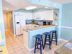 A kitchen with white cabinets, a refrigerator, a microwave, a stove, and a coffee maker. There are three blue bar stools and a glass table.