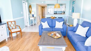 Un salon avec un canapé bleu, une chaise en bois, une table basse et, au fond, une cuisine.
