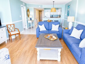 A living room with a blue sofa, a wooden chair, a coffee table, and a kitchen in the background.