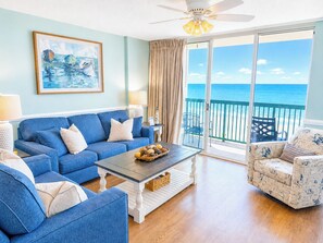 A living room with a blue sofa, a patterned armchair, a coffee table with a fruit bowl, and a painting of sailboats.