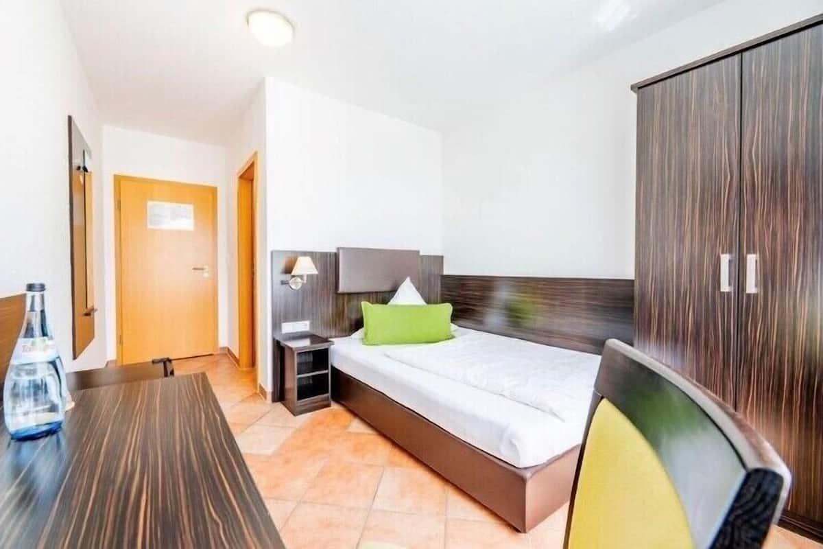 A hotel room with a bed, a nightstand, a lamp, a wardrobe, and a table with a bottle.