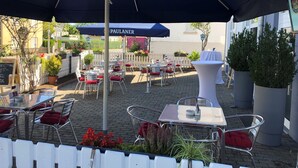 An outdoor patio area with tables and chairs, covered by umbrellas, and surrounded by potted plants and a white picket fence.