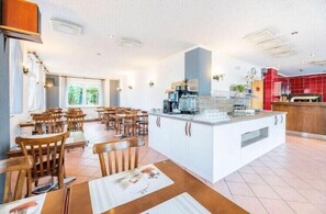 A spacious dining area with wooden tables and chairs, a central island with kitchen appliances, and a bright, well-lit interior. - My Pension (Leinfelden-Echterdingen)
