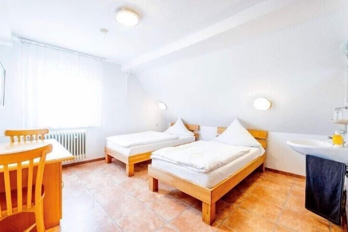A room with two single beds, a wooden table with chairs, a small sink, and a window with curtains.